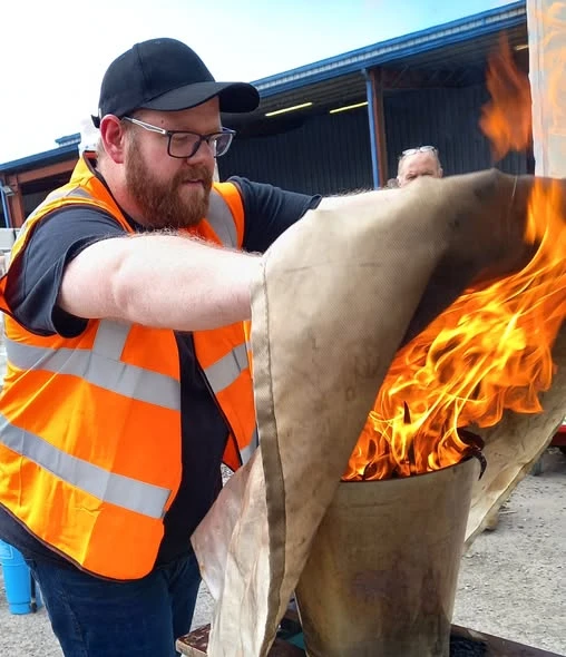 A person in a safety vest is managing a fire in a metal container, demonstrating fire safety or controlled burning techniques.