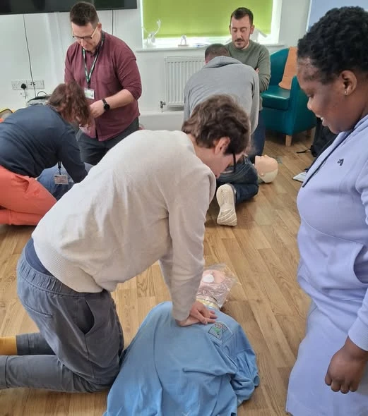 A group of people are practicing CPR on training mannequins on a wooden floor, with various individuals engaged in the activity.